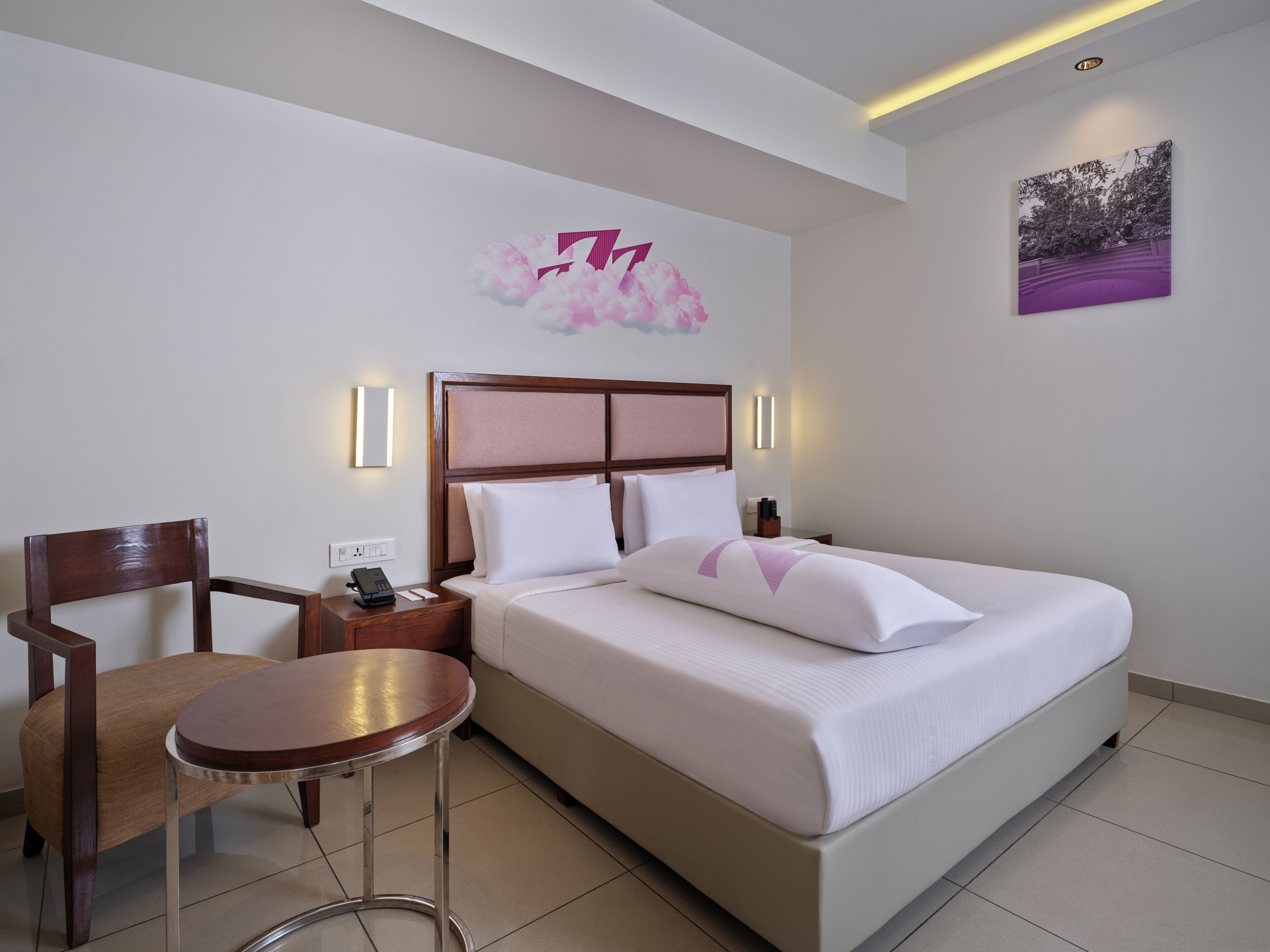 Une chambre d’hôtel comprenant un lit, une chaise, une petite table, une table de chevet, un téléphone et un luminaire fixé au mur.