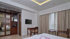 Quarto de hotel com cama, escrivaninha, cadeira, televisão, guarda-roupa e janela com cortinas.
