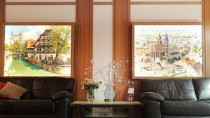 A room with two black leather sofas, a small table with a vase of flowers, and large windows with a view of a town. - Hotel am Buchwald (Esslingen)