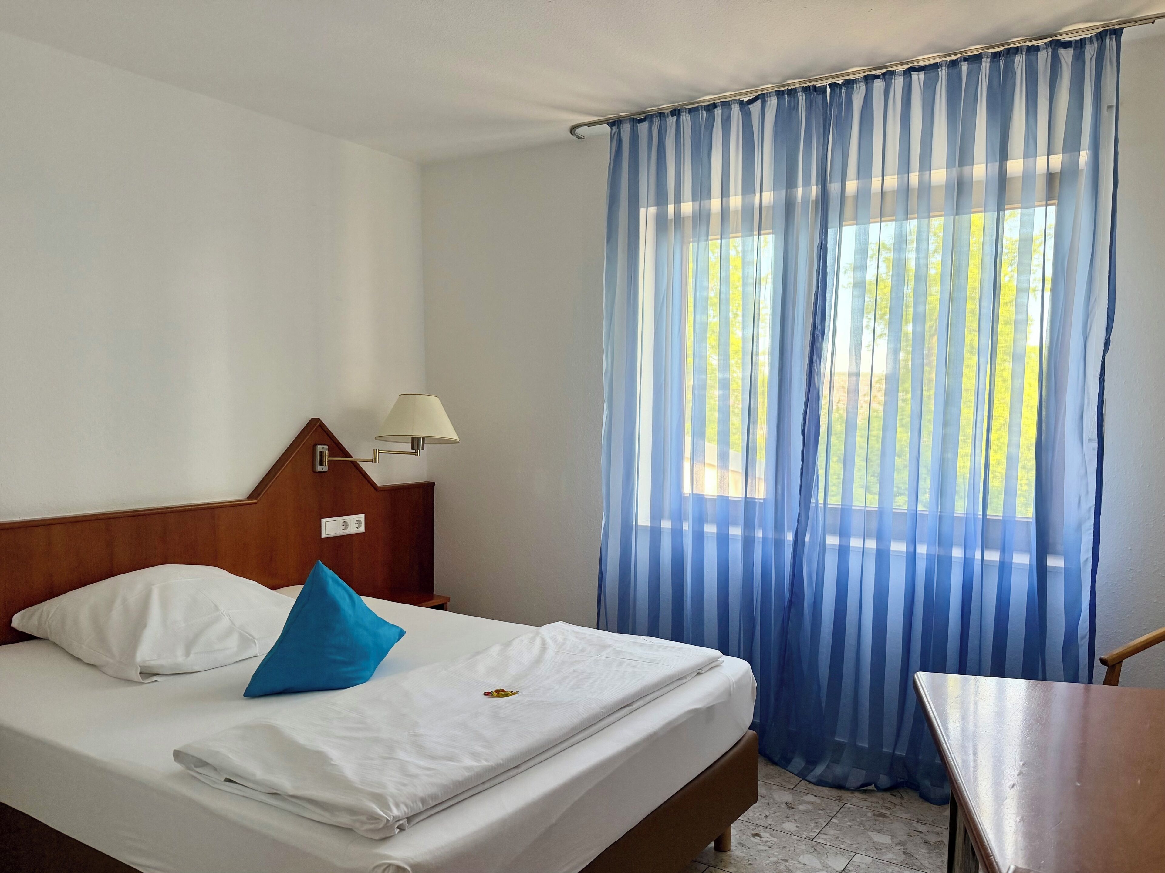Quarto de hotel com cama, mesa de cabeceira, abajur e janela com cortinas.