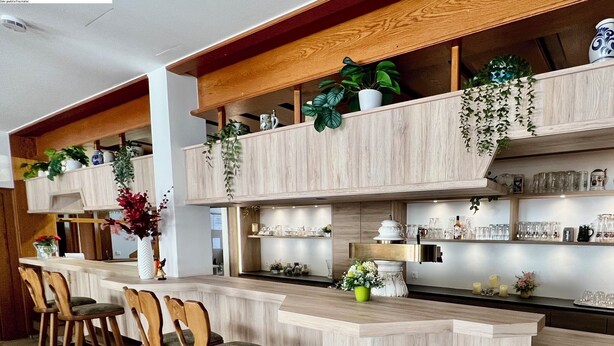 A bar area with wooden chairs, shelves with plants, and various barware. - Hotel am Buchwald (Esslingen)