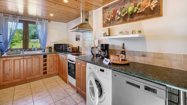 A kitchen with wooden cabinets, a washing machine, a coffee maker, and a window with curtains. - Apartment “La Poya” with Private Terrace, Garden, and Wi-Fi (Plonévez-du-Faou)