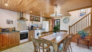 A kitchen with wooden cabinets, a dining table with chairs, a stove, a washing machine, and a clock on the wall. - Apartment “La Poya” with Private Terrace, Garden, and Wi-Fi (Plonévez-du-Faou)