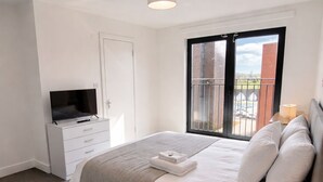 A bedroom with a bed, a TV, a desk, a window with a balcony view, and a ceiling light. - Luxury 2BR Apartment - Free parking (Slough)