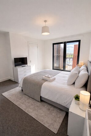 A bedroom with a bed, a TV, a desk, a window with a balcony view, and a ceiling light. - Luxury 2BR Apartment - Free parking (Slough)