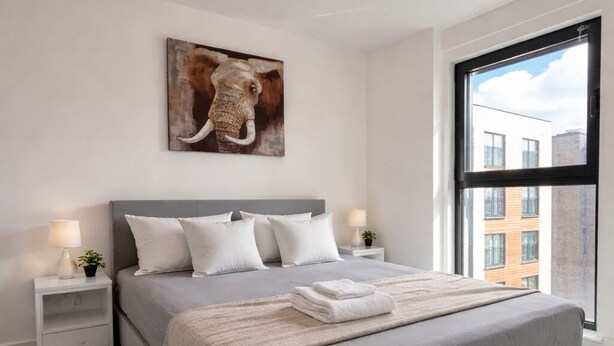 A modern bedroom with a large bed, a nightstand with a lamp, a painting on the wall, and a view of buildings through a window. - Luxury 2BR Apartment - Free parking (Slough)