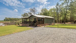 A lodge with a covered porch, a gravel driveway, and a pond in the background.