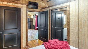 A room with wooden walls, a bed with a white mattress and a red blanket, a television mounted on the wall, and a door leading to another room.