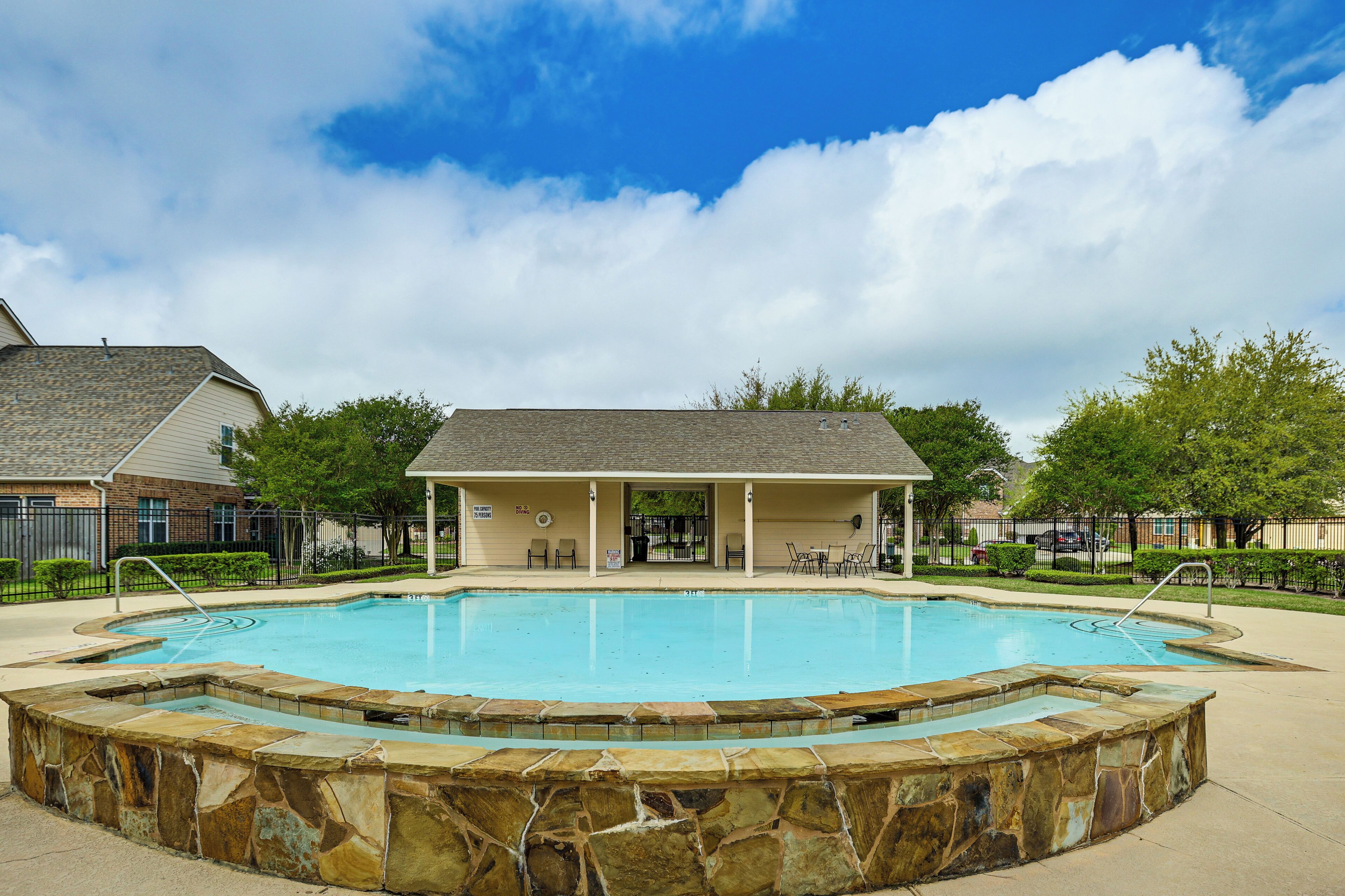A swimming pool with a stone border, a covered seating area, and a building in the background.