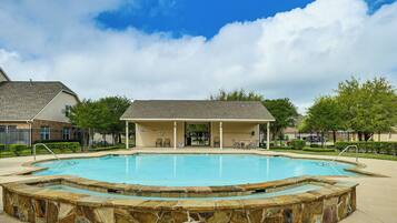 A swimming pool with a stone border, a covered seating area, and a building in the background.