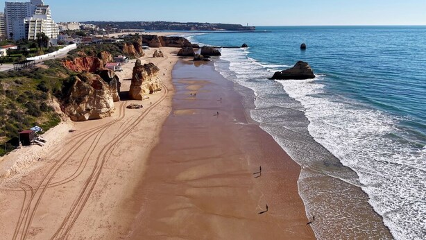 Uma praia costeira com falésias rochosas, uma faixa de areia e edifícios ao fundo.