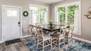 A dining area with a round table, six chairs, a vase with flowers, and a view through large windows. - Greenwood Group Retreat w/ Large Private Dock! (Greenwood)
