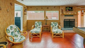 Una habitación con mesa de billar, dos sillones de mimbre con estampado tropical, una mesita con lámpara y una chimenea de ladrillo.