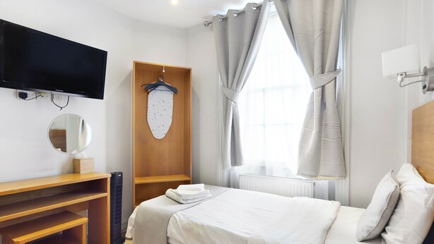 Ein ordentlich eingerichtetes Hotelzimmer mit einem Bett, einem an der Wand montierten Fernseher, einem Holztisch, einem Spiegel und einem Fenster mit Vorhängen.