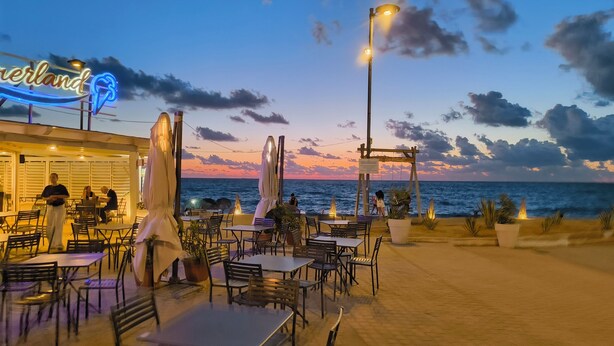 Restaurante à beira-mar com mesas ao ar livre, um letreiro de neon e vista para o oceano.