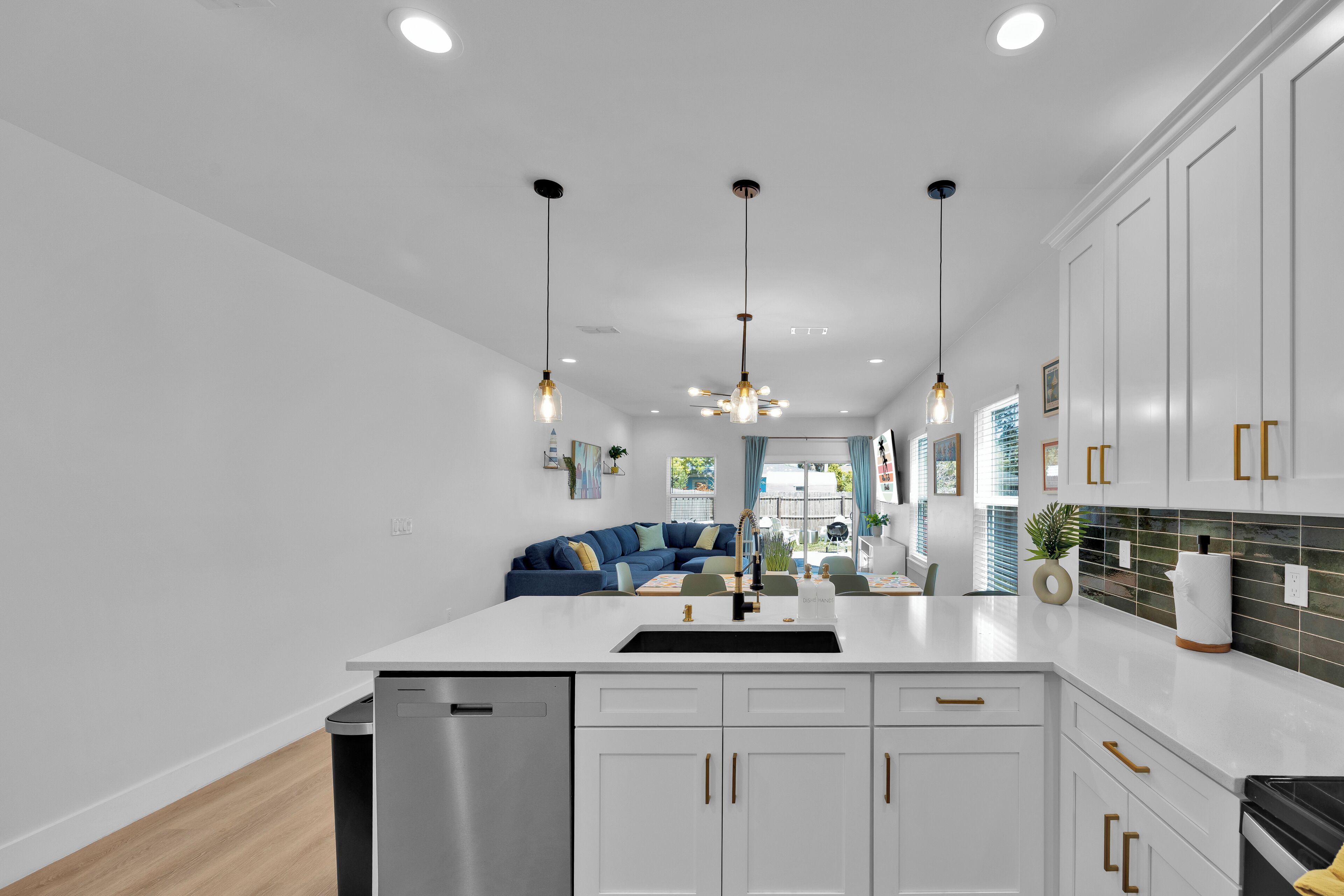 A modern kitchen with white cabinets, a stainless steel dishwasher, pendant lighting, and a view into the living area.