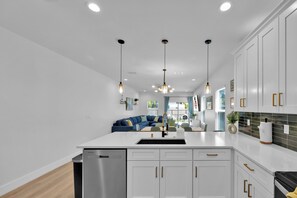 A modern kitchen with white cabinets, a stainless steel dishwasher, pendant lighting, and a view into the living area. - Beach Home | Hot tub | Bikes & E-scooters | Game Room (Jacksonville Beach)