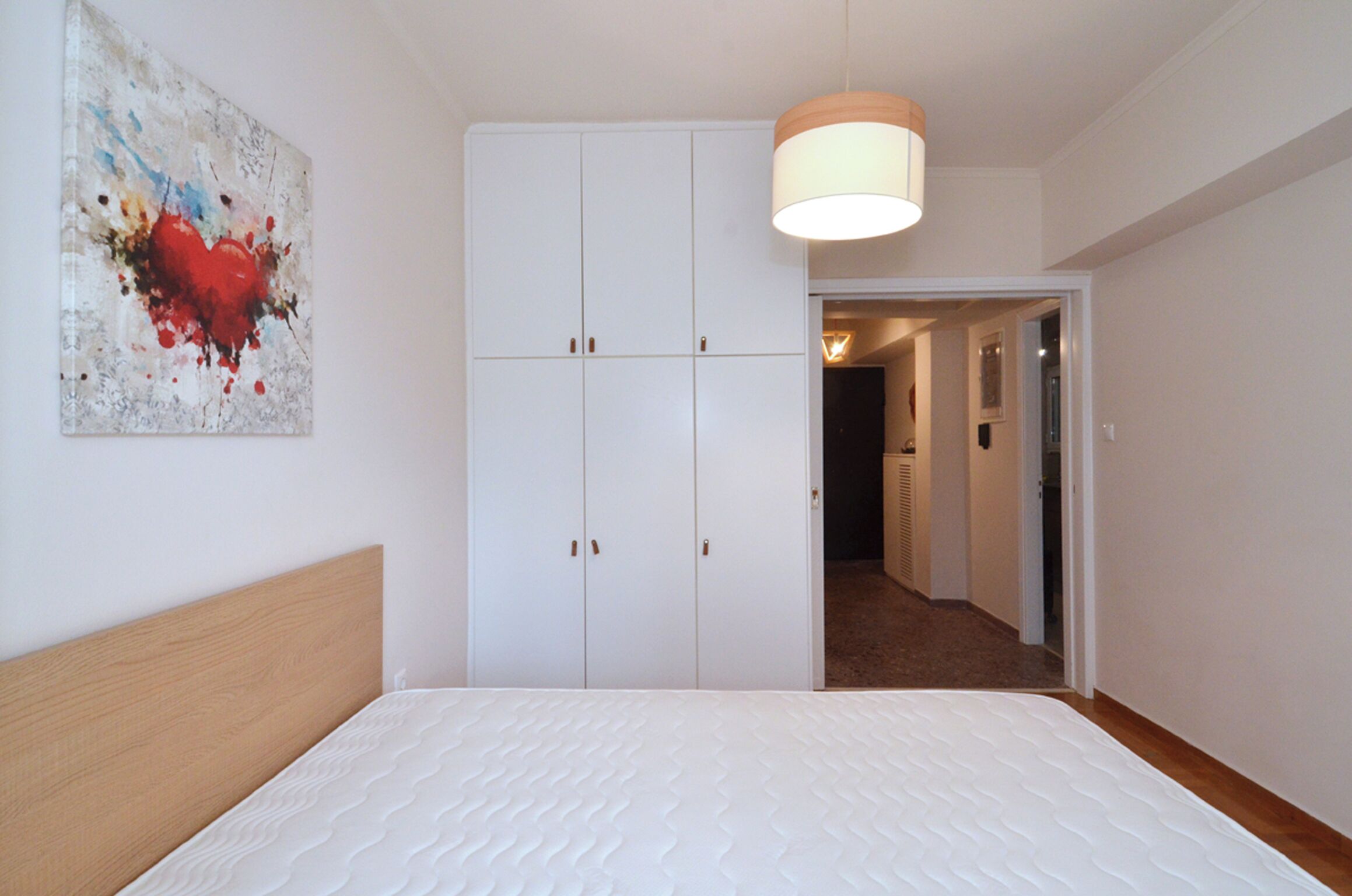 A bedroom with a bed, a white wardrobe, a painting on the wall, and a hallway leading to another room.