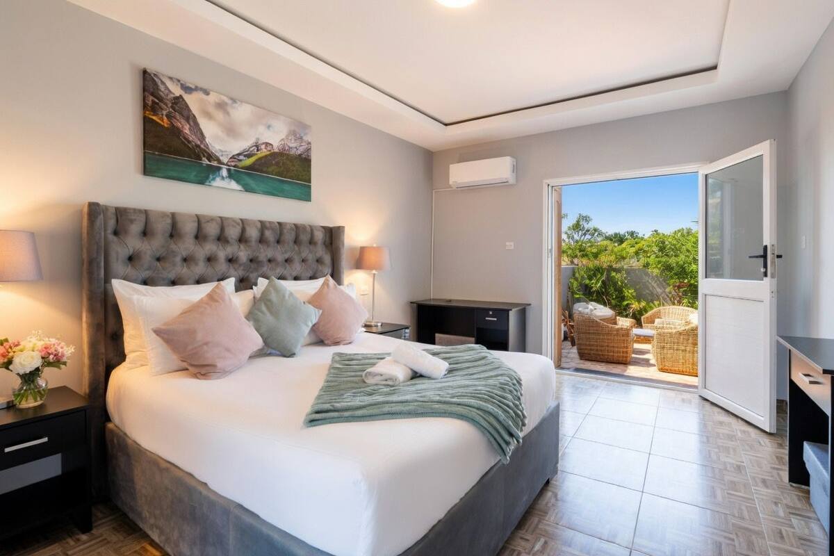 Ein Schlafzimmer mit einem großen Bett, einem Gemälde darüber, einem Nachttisch mit Lampe, einem Schreibtisch und Blick ins Freie durch eine offene Tür.
