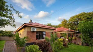 Une maison à deux étages, avec un toit rouge, une façade en briques et un jardin où poussent différentes espèces de plantes.