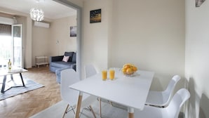 A dining area with a white table, chairs, and a fruit bowl. There is a sofa and a lamp in the background. - Ano Patisia A Fully Renovated Penthouse (Athens)