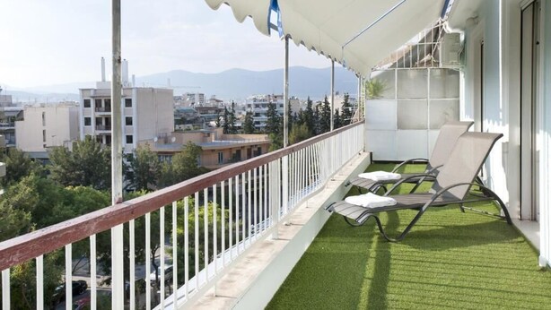 A balcony with artificial grass, two lounge chairs, a white railing, and a view of buildings and mountains. - Ano Patisia A Fully Renovated Penthouse (Athens)
