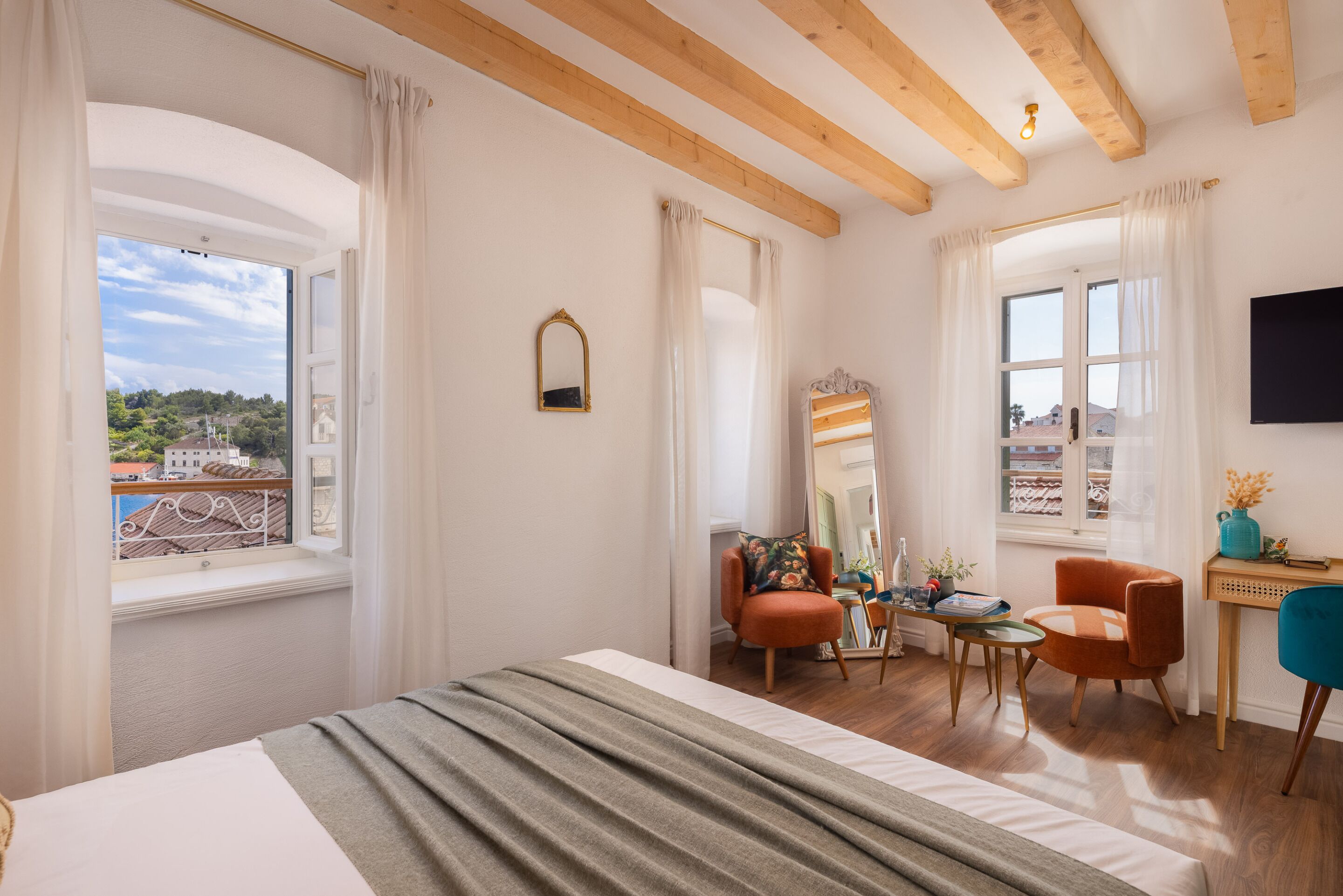 Une chambre à coucher comprenant un lit, un bureau, deux chaises, un miroir et une fenêtre donnant sur des immeubles.