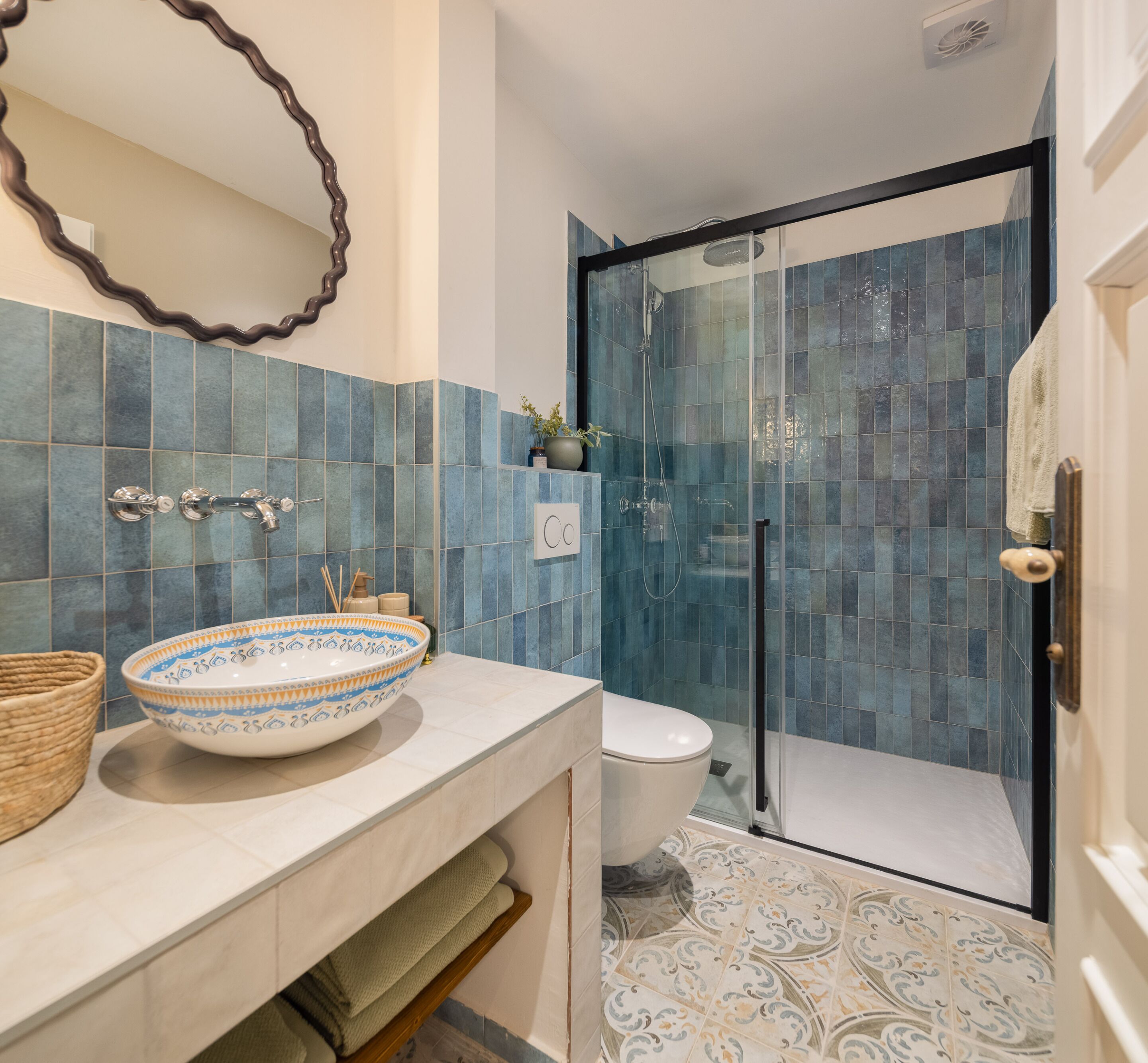 Une salle de bain dotée d’une douche à parois de verre, d’un lavabo blanc agrémenté d’un récipient décoratif et d’un miroir dont le cadre est ondulé.