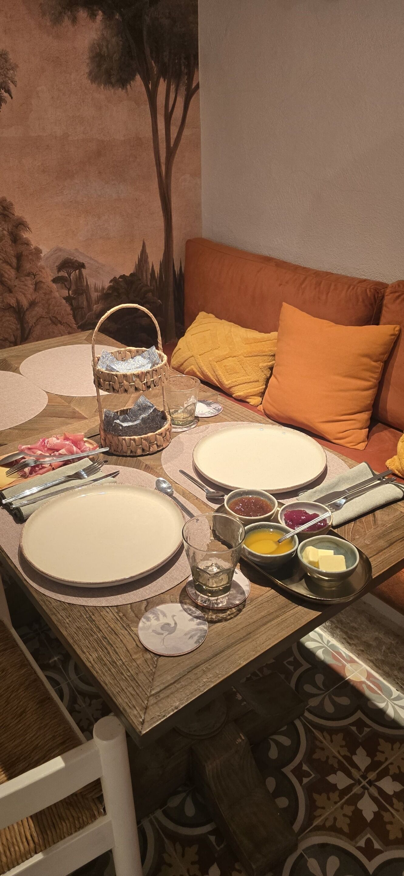 Une table à manger dressée avec des assiettes, des bols et des couverts, un panier en osier et un verre d’eau.