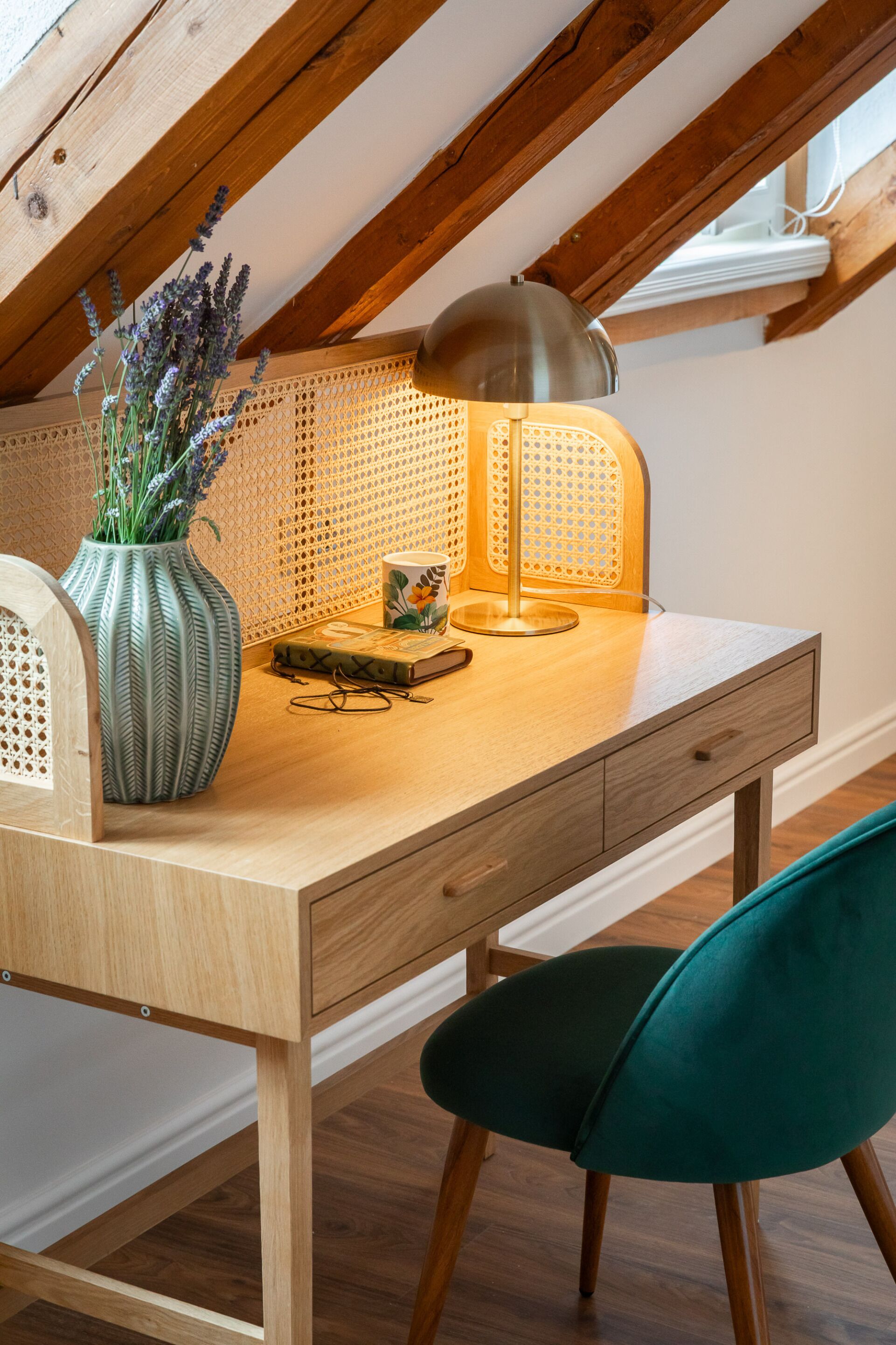 A wooden desk with a lamp, a vase with flowers, and a chair in a room with wooden beams and a sloped ceiling.