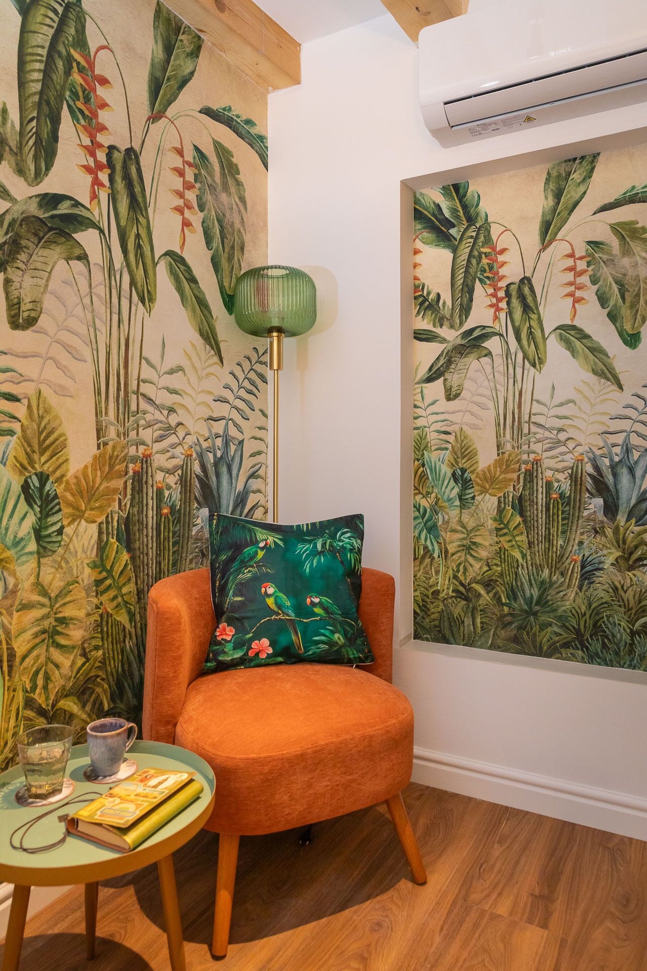 A room with a tropical-themed mural, a single orange chair, a small table with a book and a cup, and a floor lamp.