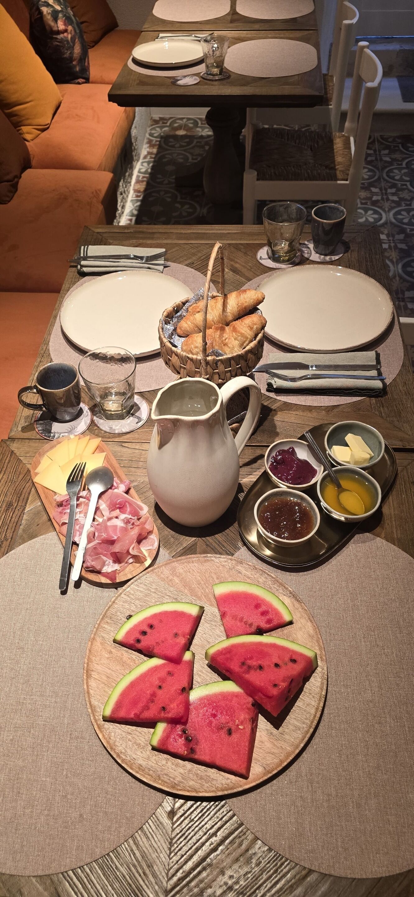 A dining table set with plates, cutlery, a pitcher, a basket of croissants, bowls of jam and butter, a plate of watermelon, and a glass of water.