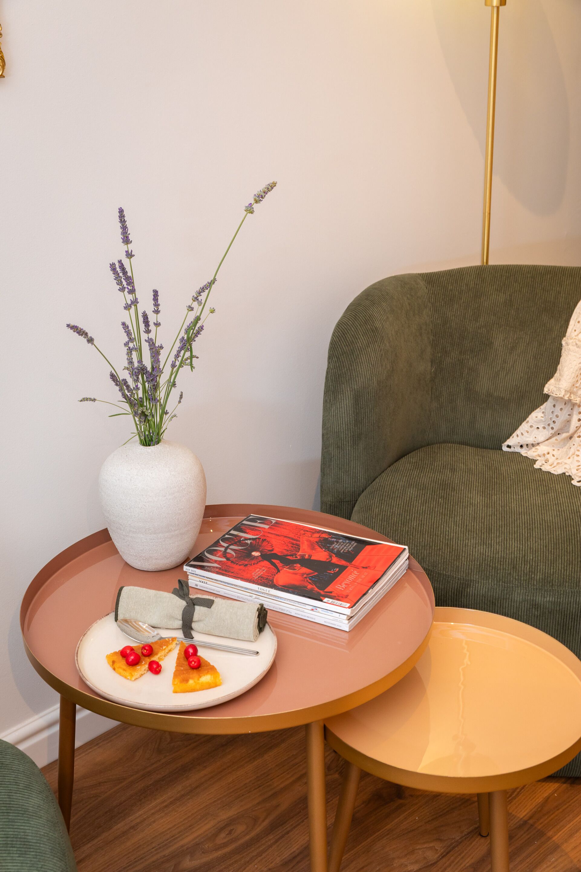A room with a round wooden table, a green sofa, a white vase with lavender, a plate with food, and a magazine.