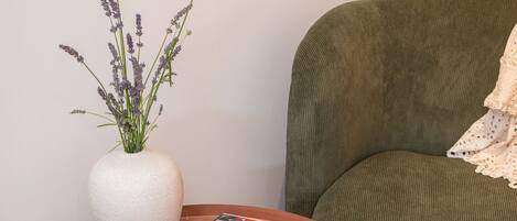 A room with a round wooden table, a green sofa, a white vase with lavender, a plate with food, and a magazine.