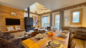 Ein gemütliches Blockhaus-Interieur mit Essbereich, Kamin, Bücherregal und Fenster mit Blick auf Bäume.