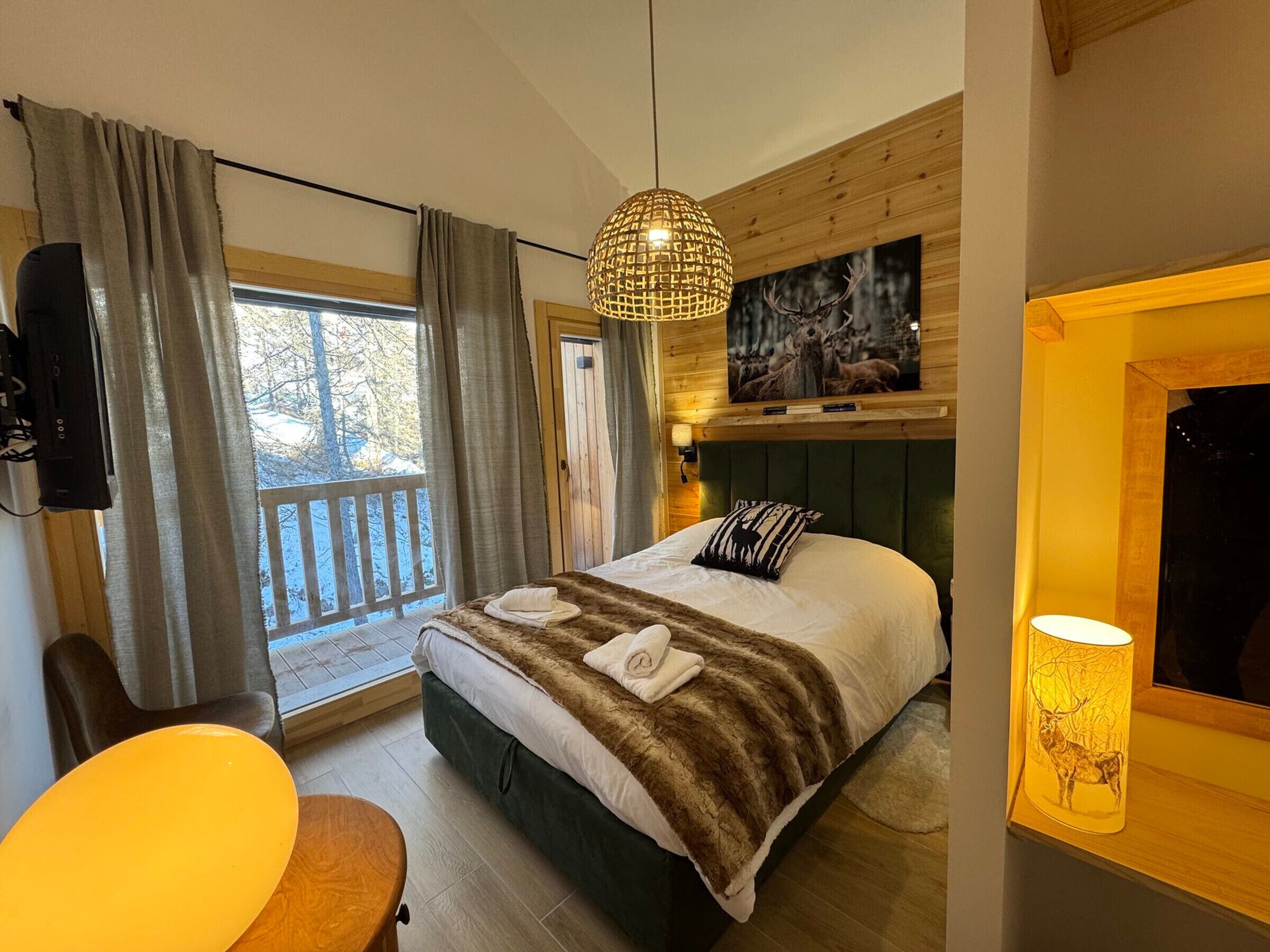 A bedroom with a bed, a wooden headboard, a window with curtains, a mounted TV, a table, and a wall-mounted lamp.