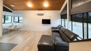 Uma sala de estar moderna com teto de madeira, sofá de couro preto, área de jantar com mesa e cadeiras, e uma janela grande com vista para o exterior.