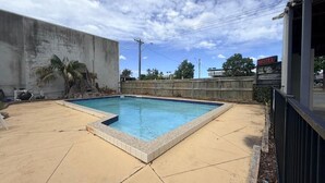 Piscina com borda azulejada, um pátio de concreto, cerca de madeira e um edifício com a placa 'HOTEL'.