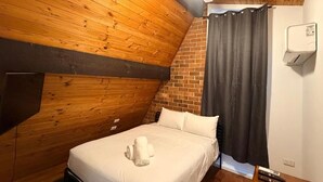 Quarto com vigas de madeira no teto, cama com roupa de cama branca, cortina preta e ventilador de teto.