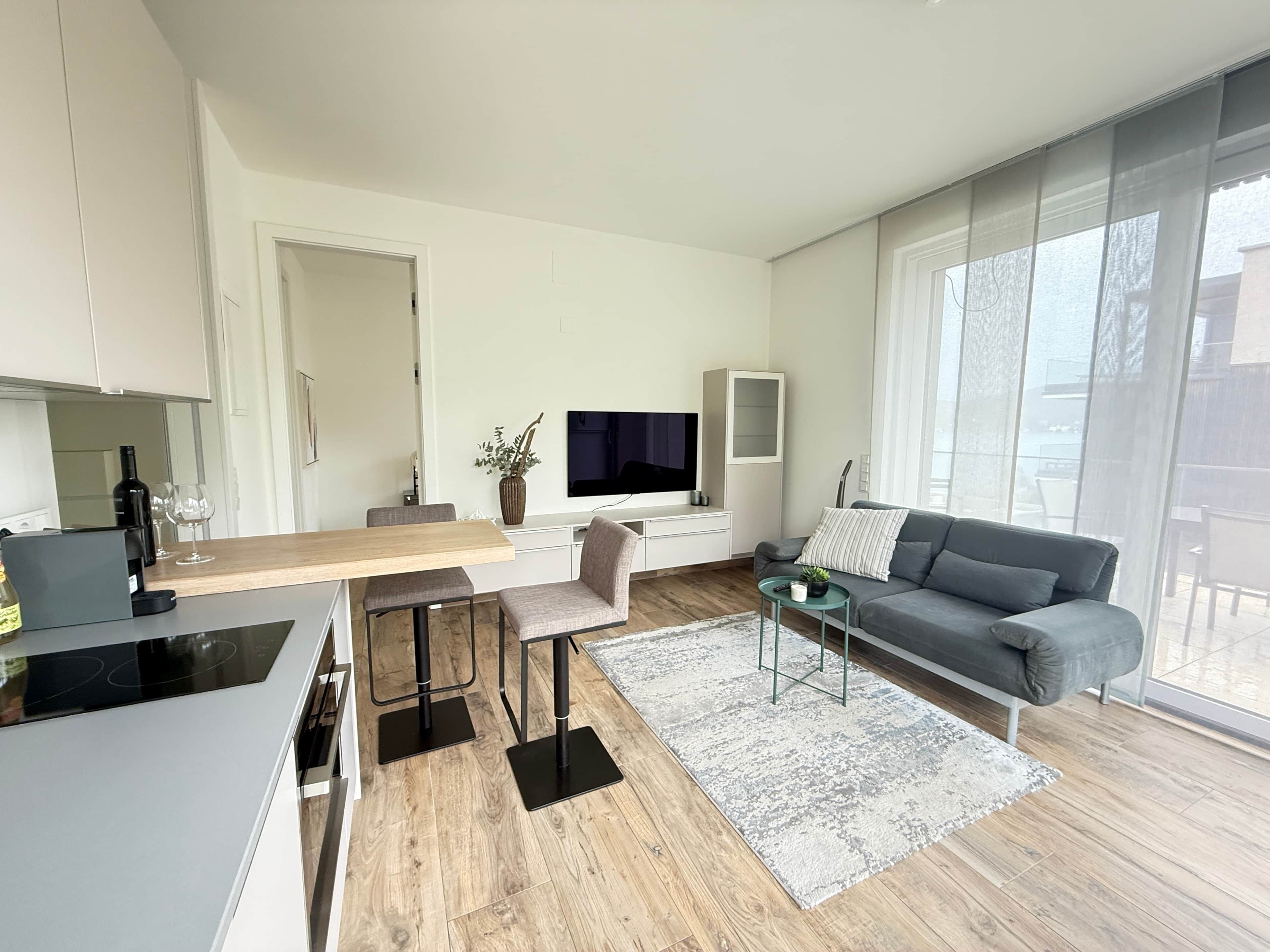 A modern living room with a grey sofa, a TV, a dining table with chairs, and a kitchen area with a stove.
