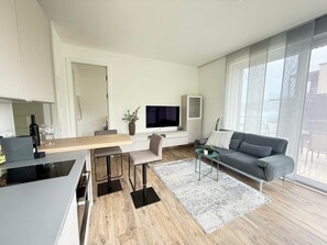 A modern living room with a grey sofa, a TV, a dining table with chairs, and a kitchen area with a stove. - Hermitage Apartment A09 - Lake view & access (Maria Wörth)
