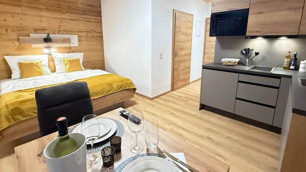 Una habitación compacta con cama, una mesa de comedor para dos personas, kitchenette y techo de madera.