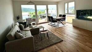 Amplia sala de estar con sofá, mesa de centro y mesa de comedor, ventanas grandes con vistas al exterior y un televisor de pantalla plana montado en la pared.