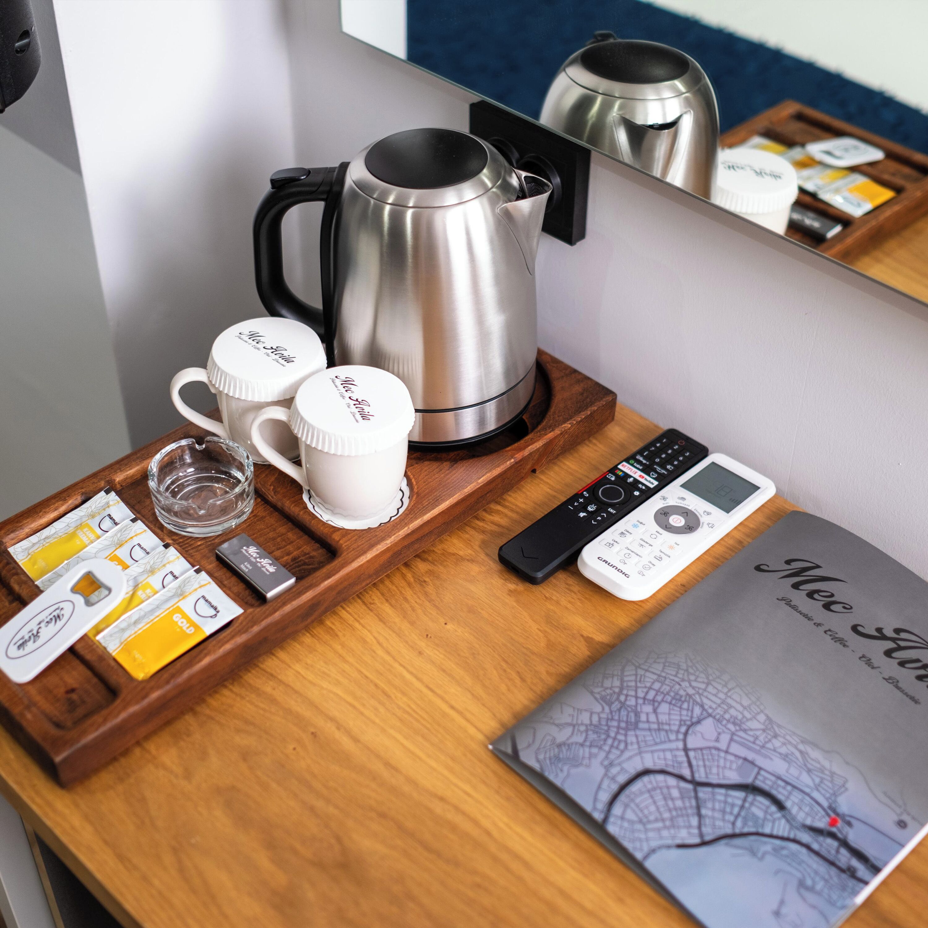 A hotel room with a wooden tray holding a coffee maker, two white mugs, a remote control, a smartphone, and a booklet.