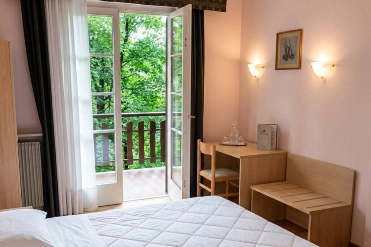 Habitación de hotel con cama, escritorio, silla, ventana con vistas a vegetación y una lámpara fijada a la pared.