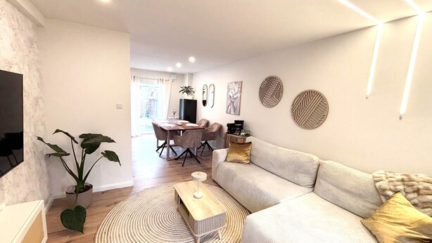 A modern living room with a sofa, a coffee table, a dining area with chairs, and a TV mounted on the wall. - Holiday Home 'Kokorey' with Private Terrace, Garden and Wi-Fi (Rellingen)
