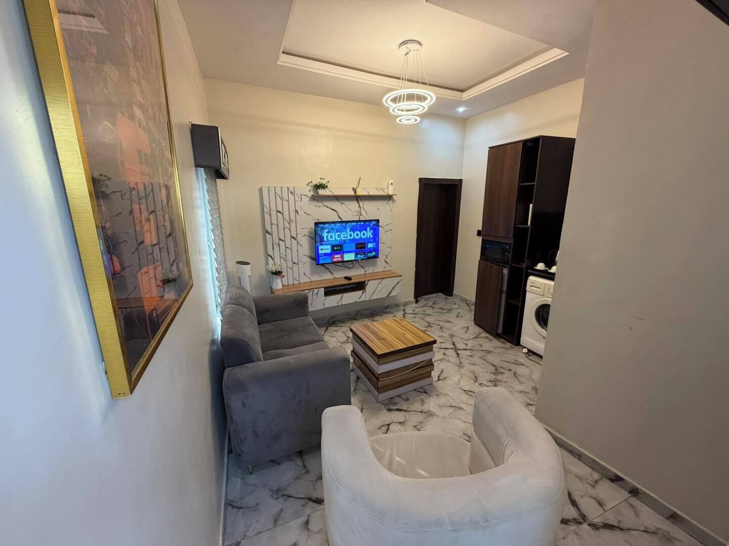 A modern living room with a sofa, a white armchair, a coffee table, a TV mounted on the wall, and a kitchen area with appliances.