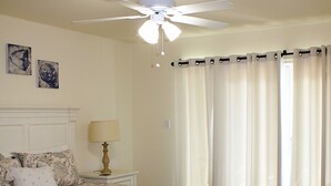 Ein Schlafzimmer mit Bett, Nachttisch, Lampe, Deckenventilator und Vorhängen.