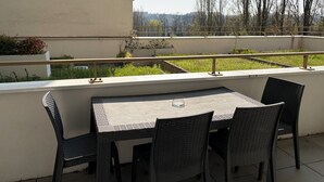 Un balcón con mesa y sillas, barandilla y vista a árboles y un paisaje lejano.