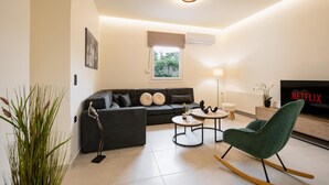 A modern living room with a sofa, two side tables, a green rocking chair, a TV, and a window with blinds. - Apartment 'Zoelgrek Luxury Suite 1' with Private Terrace, Wi-Fi and Air Conditioning (Iraklio)